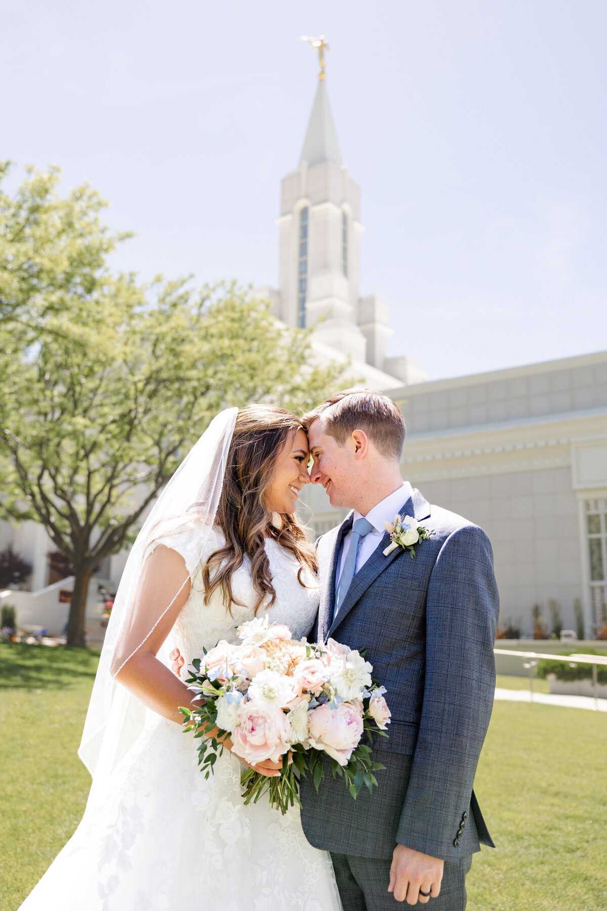 A newlywed couple standing on a lush green lawn in front of the Bountiful Utah Temple, gazing lovingly into each other’s eyes. The bride holds a bouquet of soft pastel flowers while wearing a lace wedding gown and veil, and the groom wears a navy blue suit with a light blue tie.