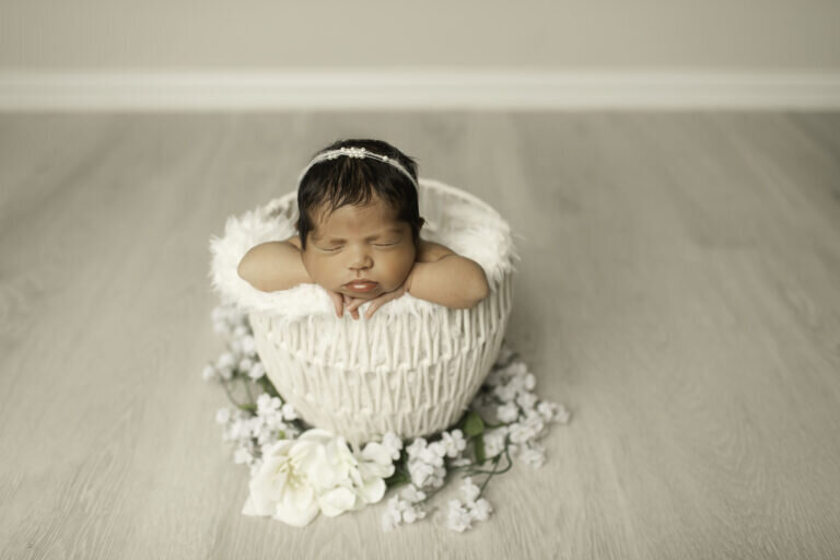Newborn baby girl peacefully posed with her hands under her chin, resting in a white wicker basket, creating a soft and delicate portrait