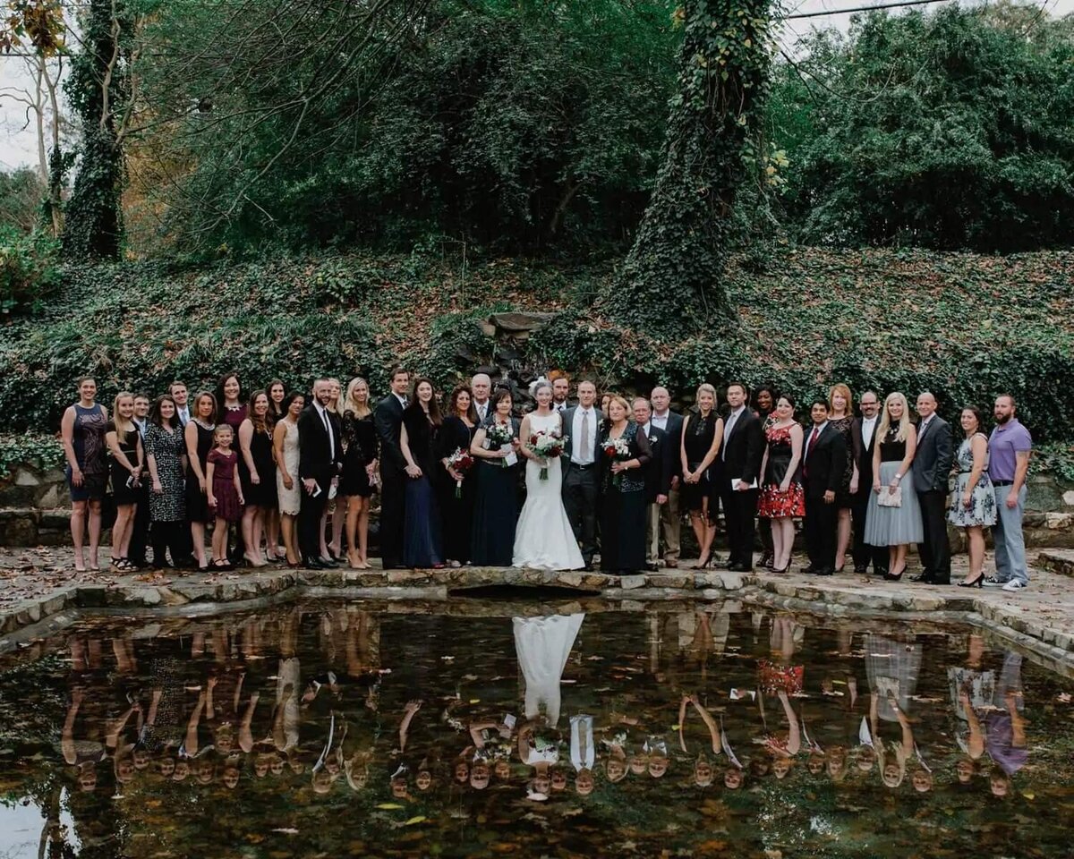A bride and groom and their wedding party standing behind a small pond.