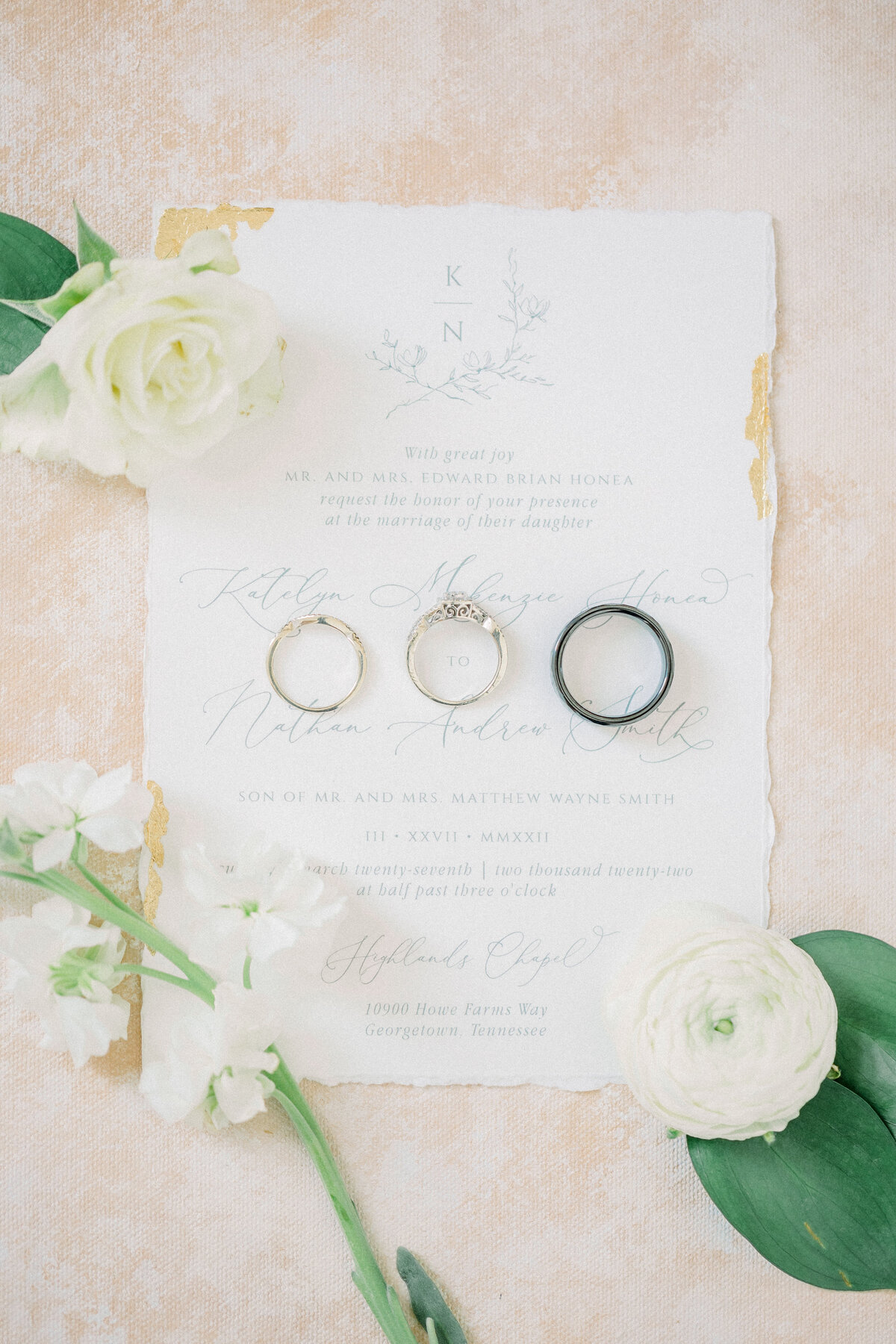 The bride's and groom's rings on their wedding invitation surrounded by white florals and greenery