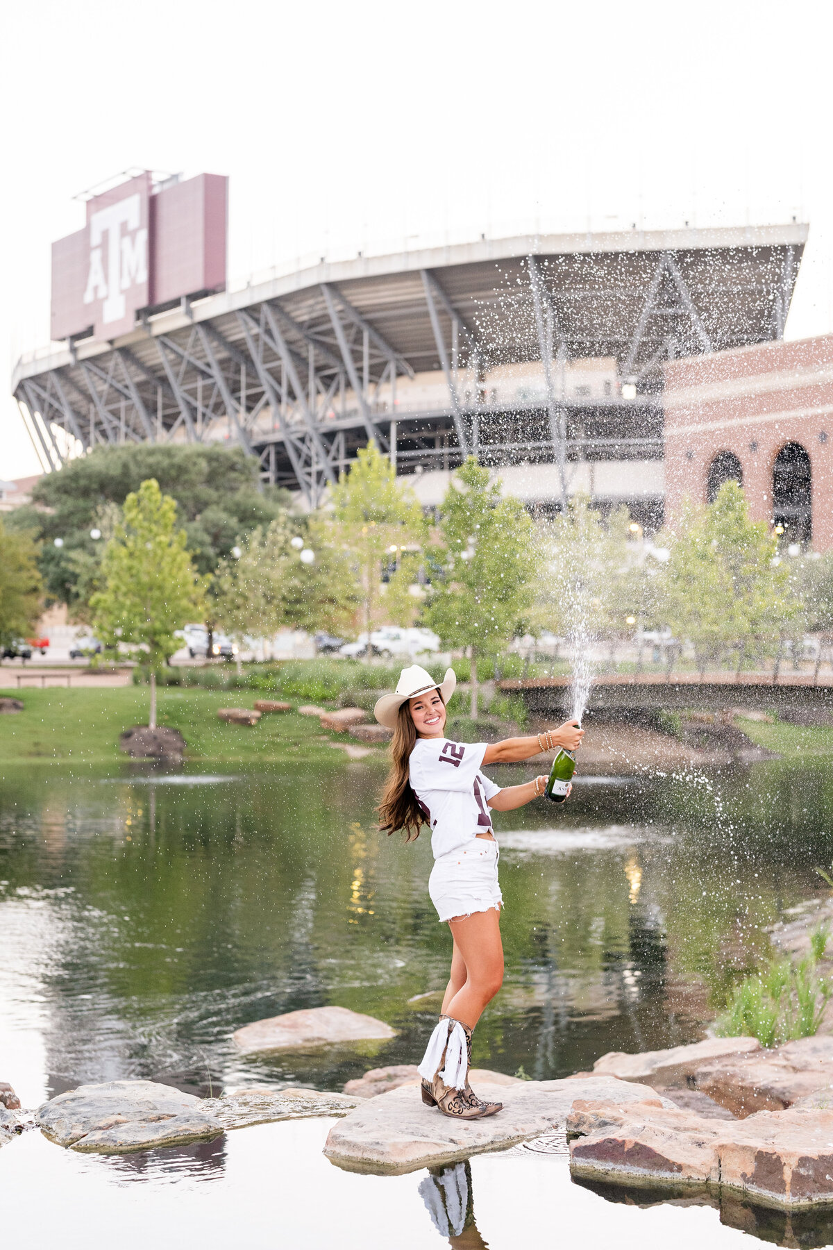 Texas A&M senior girl spraying champagne while standing on rocks in Aggie Park with Kyle Field in the background and wearing cowboy hat, white jersey and boots