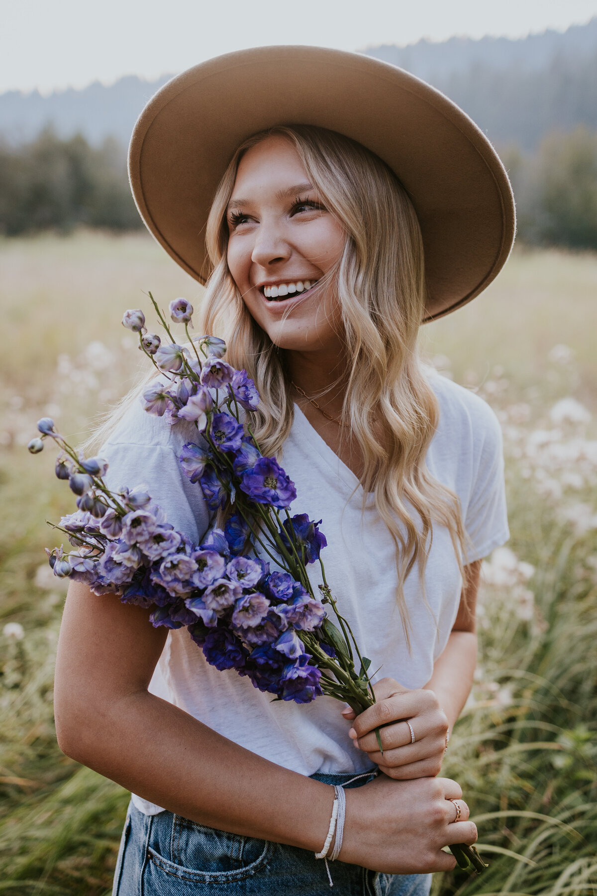 Young woman wearing beige fedora smiles to the left of camera while holding stock of purple flowers.