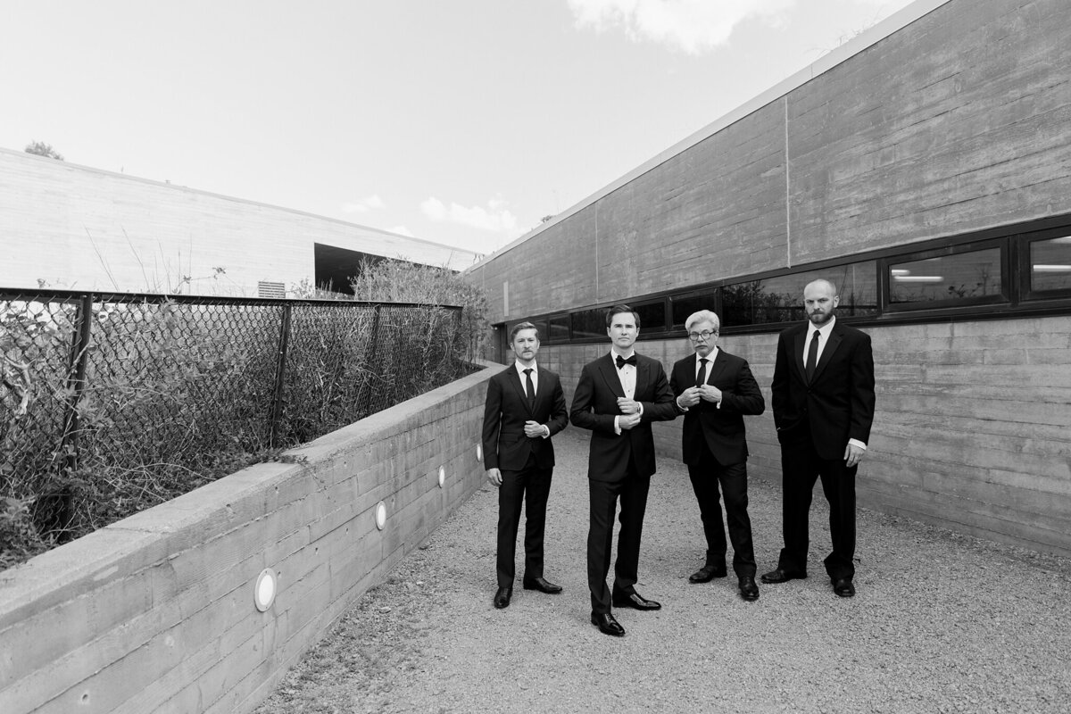 A black and white portrait of a groom and his three groomsmen on his wedding day at the Trinity River Audubon Center in Dallas, Texas. The groom is wearing a black tuxedo with a bowtie while his groomsmen all wear black suits with ties. They're all standing together in a "GQ" style of posing.