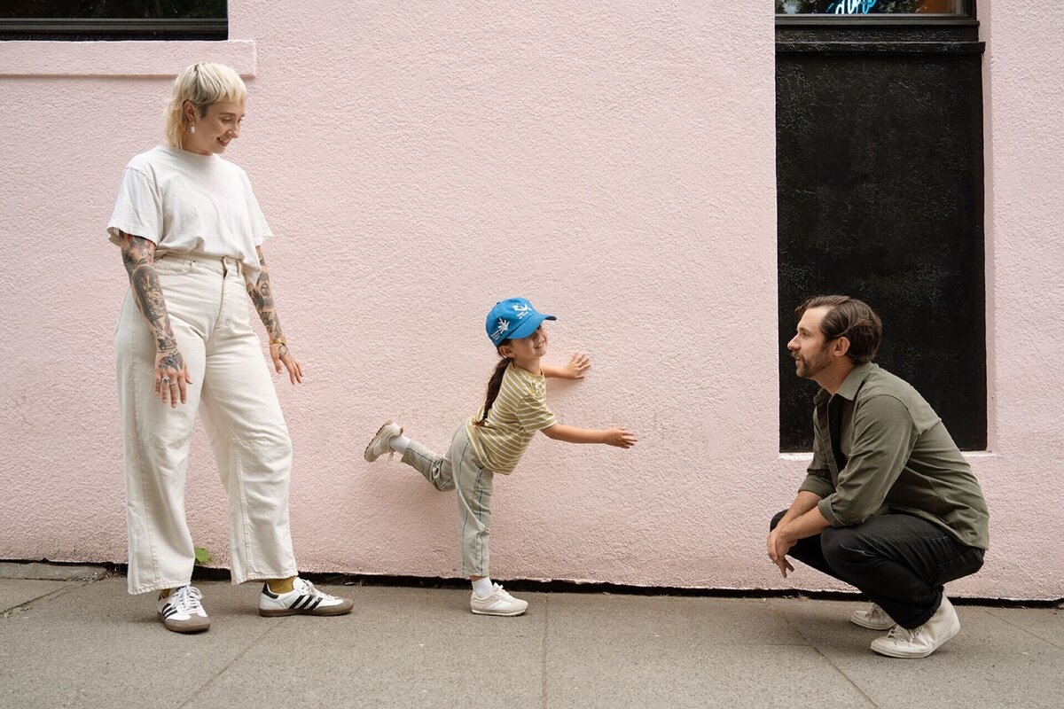 little boy wearing baseball cap playing on pink wall with male and female during family photos