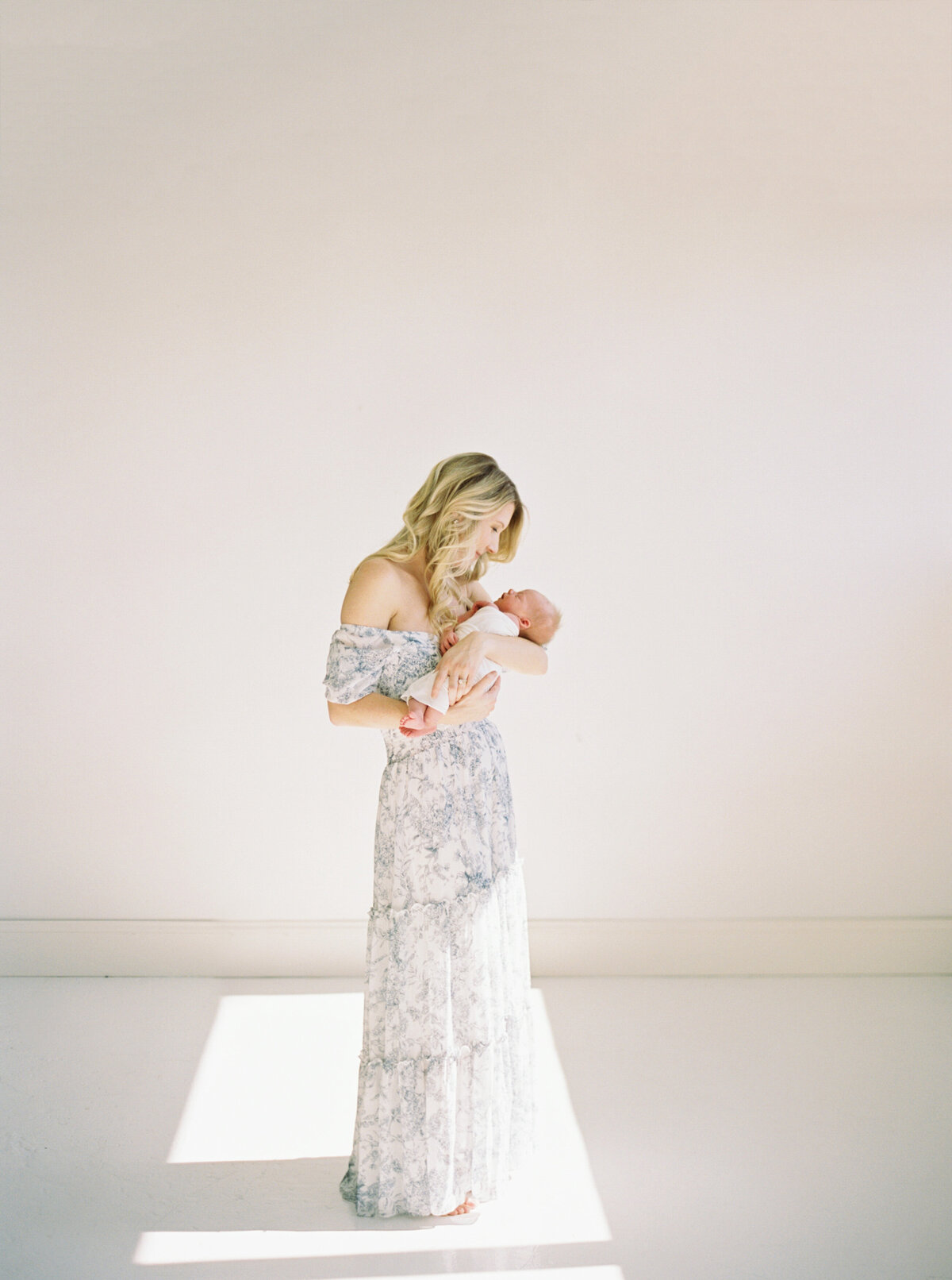 mother in blue floral dress holding newborn baby in white studio taken by newborn photography milwaukee Talia Laird Photography