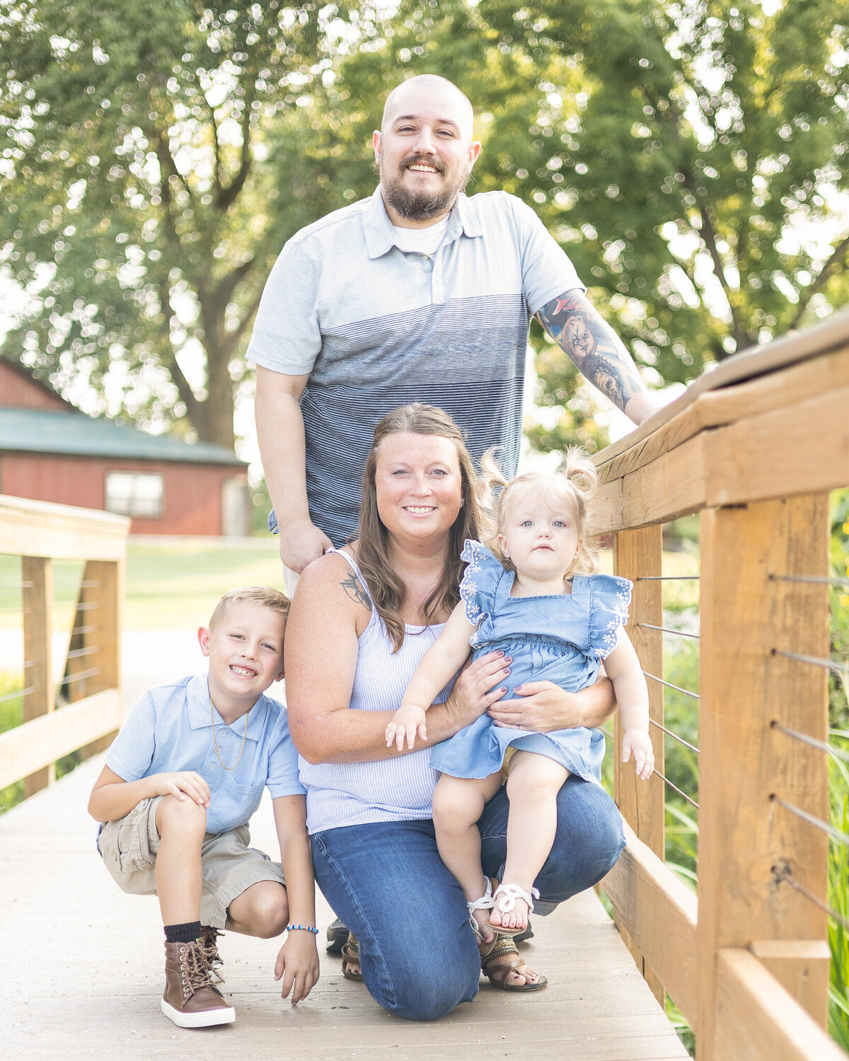 Family of found on a bridge.  Dad is standing behind family wearing a short sleeve blue shirt smiling at the camera.  Mom is knelt down with young daughter on her lap, smiling at the camera, wearing a white sleeveless shirt and blue jeans.  Sons is next to mom wearing a light blue partial button up shirt with khaki shorts, smiling at the camera, daughter has blonde hair in pigtails, wearing a light blue dress, white sandals, and looking at the camera 