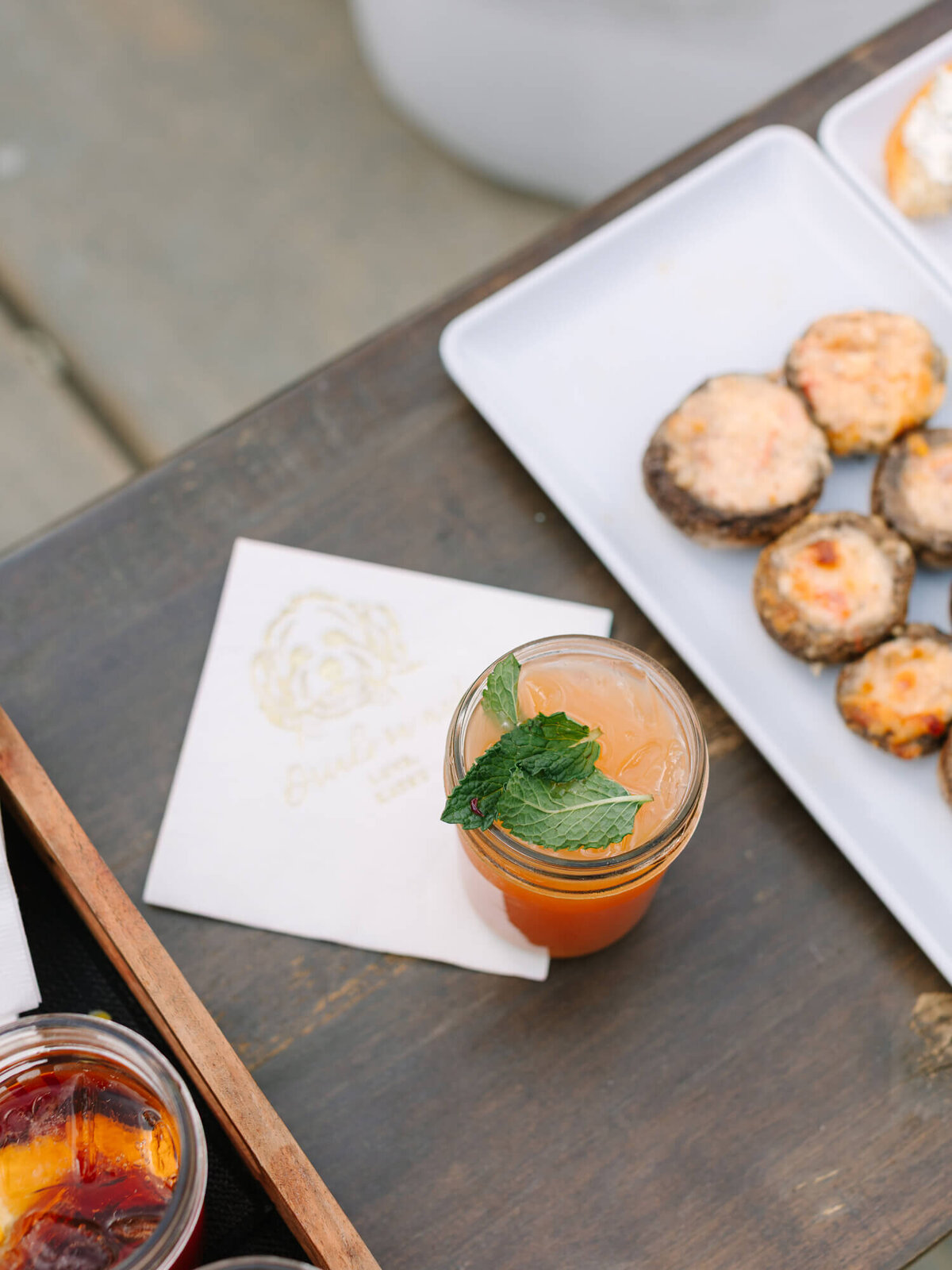 A refreshing orange drink with mint leaves on a wooden table, beside a napkin and a white tray of baked mushrooms with cheese. Cozy, inviting setting.