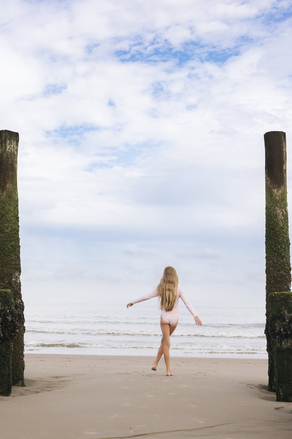 A young girl in a long-sleeved pink swimsuit by Swim Essentials walking toward the sea between two mossy wooden poles on a cloudy beach day.