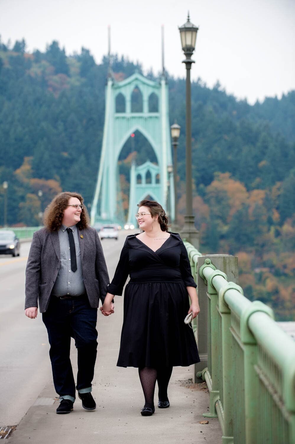 a bride in a black wedding dress holds hands and walks across the st johns bridge