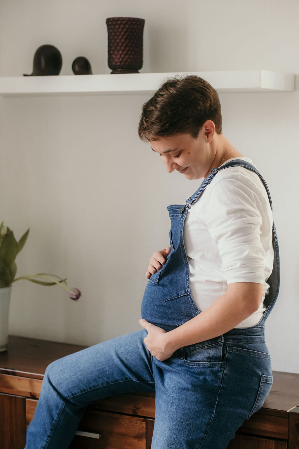 The pregnant woman sits on a sideboard holding her baby bump.