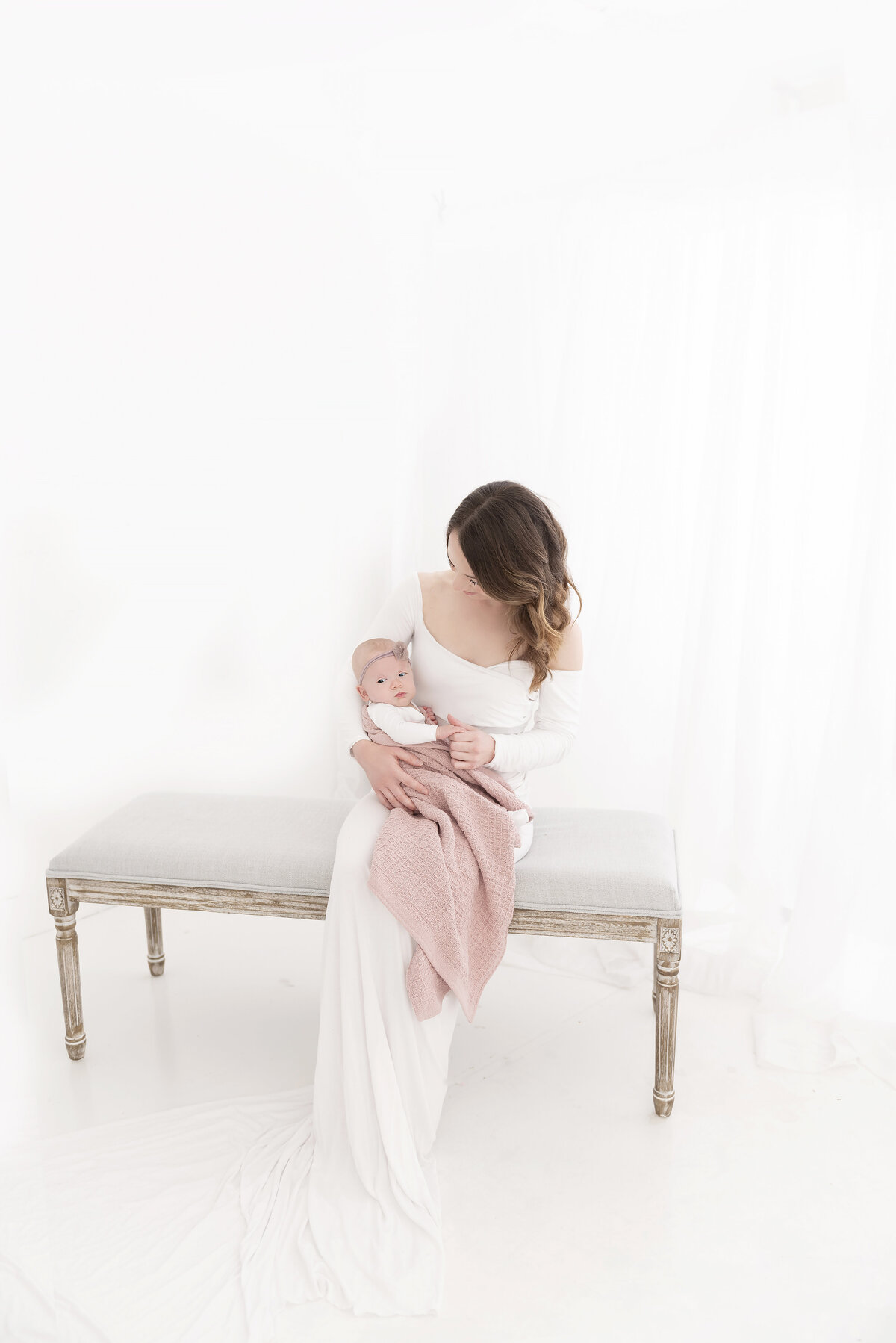 A new mother in a white dress sits on a bench in a white studio playing with her newborn baby wrapped in a pink blanket