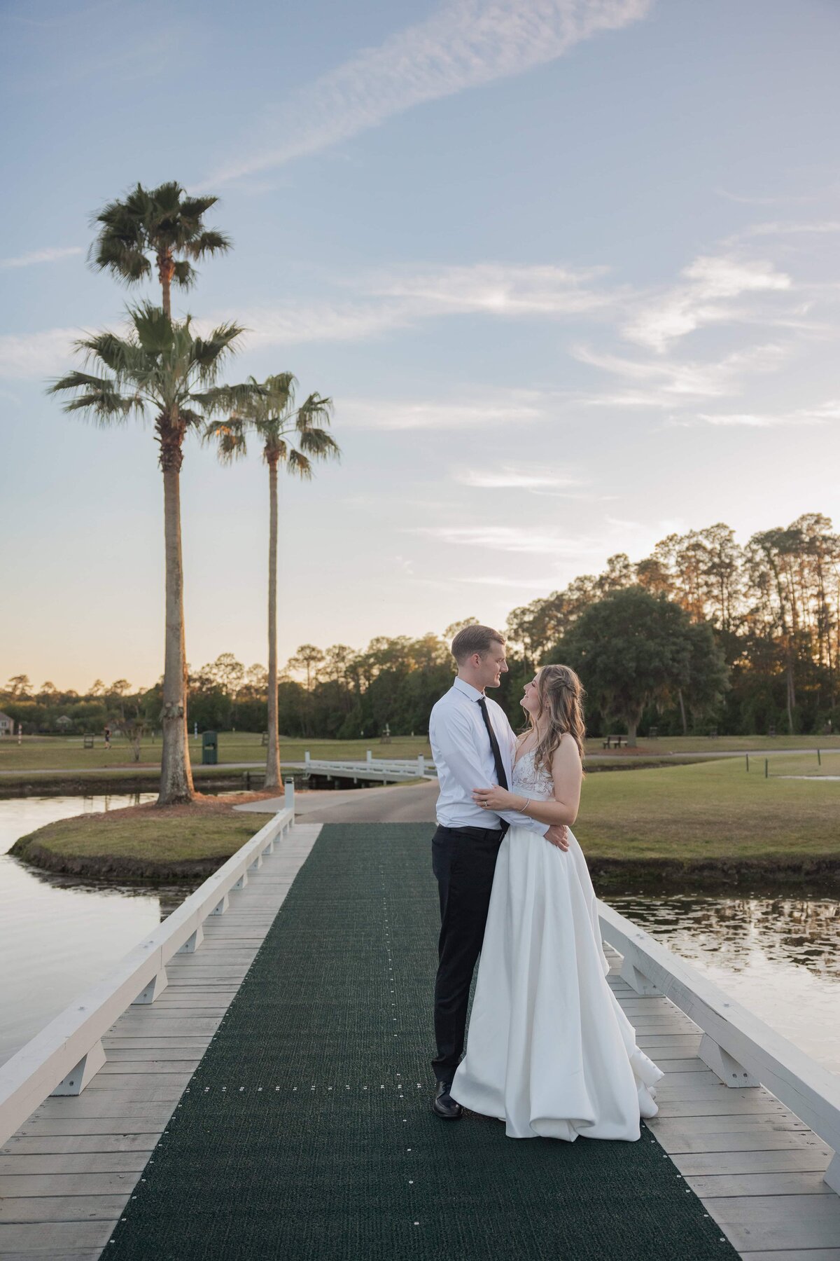 Professional wedding photography at sunset featuring a bride and groom embracing on a dock surrounded by palm trees and water. Captured during golden hour at a Jacksonville golf course by Phavy Photography, specializing in timeless and true-to-color wedding imagery.