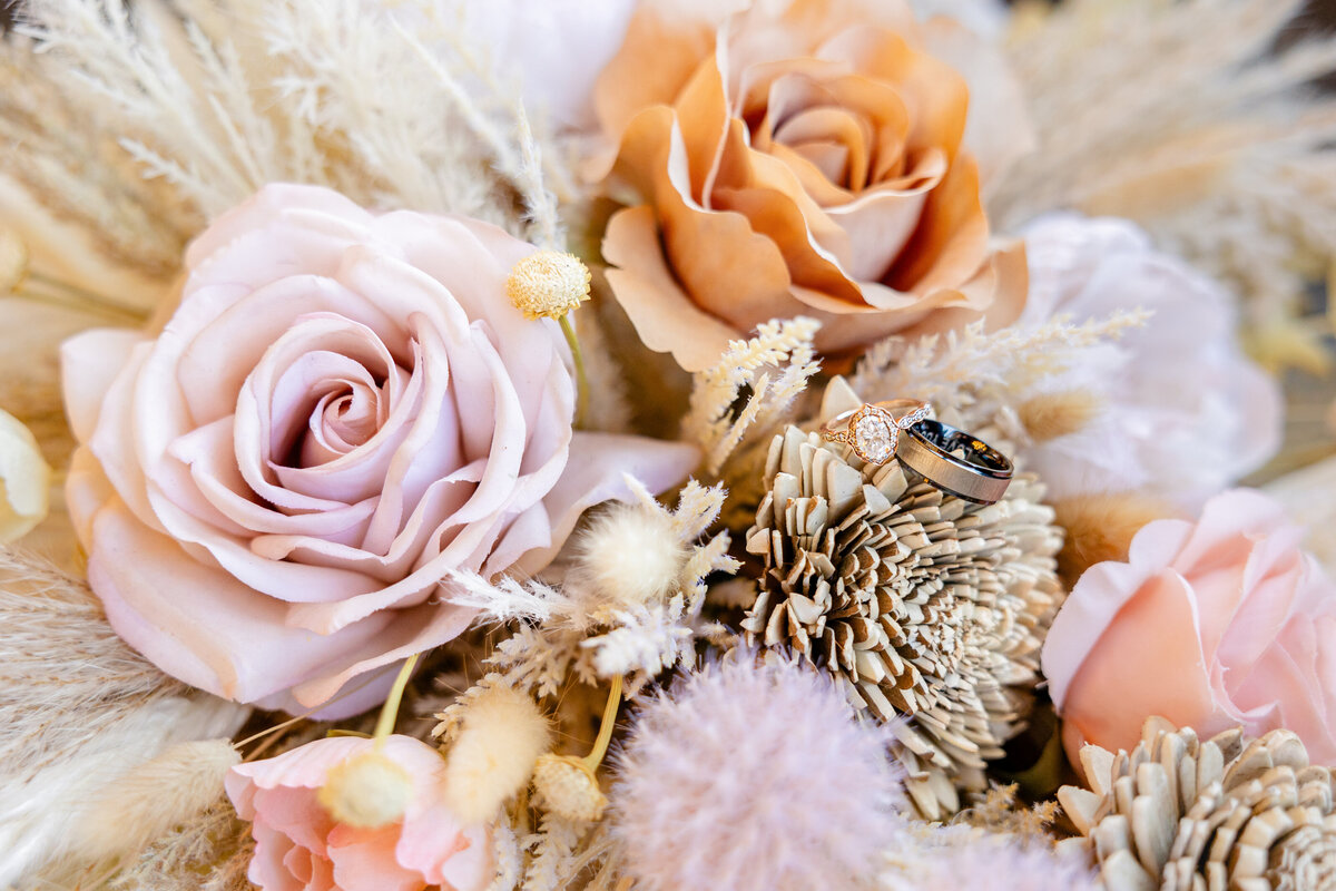 A stunning close-up of a rose gold engagement ring and a groom's wedding band nestled among pastel and earthy-toned flowers. The intricate floral arrangement adds a timeless, romantic touch.