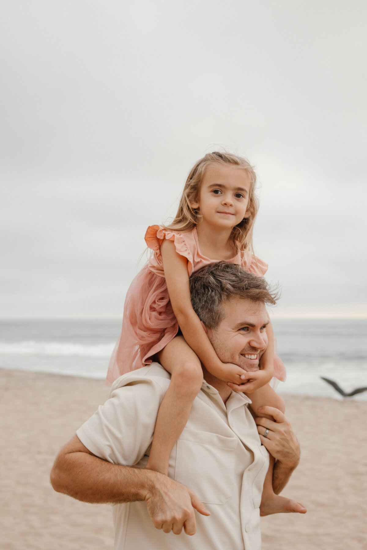 daughter rides on dad's shoulder's while smiling at the camera with the ocean in the background
