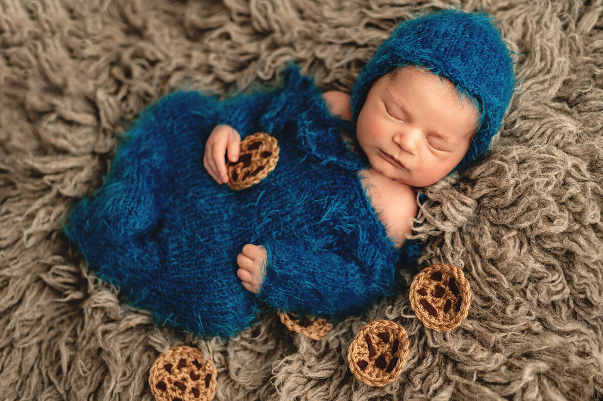 New baby boy asleep in blue hat and sleeper on grey fur surrounded by cookies at in studio photography session.
