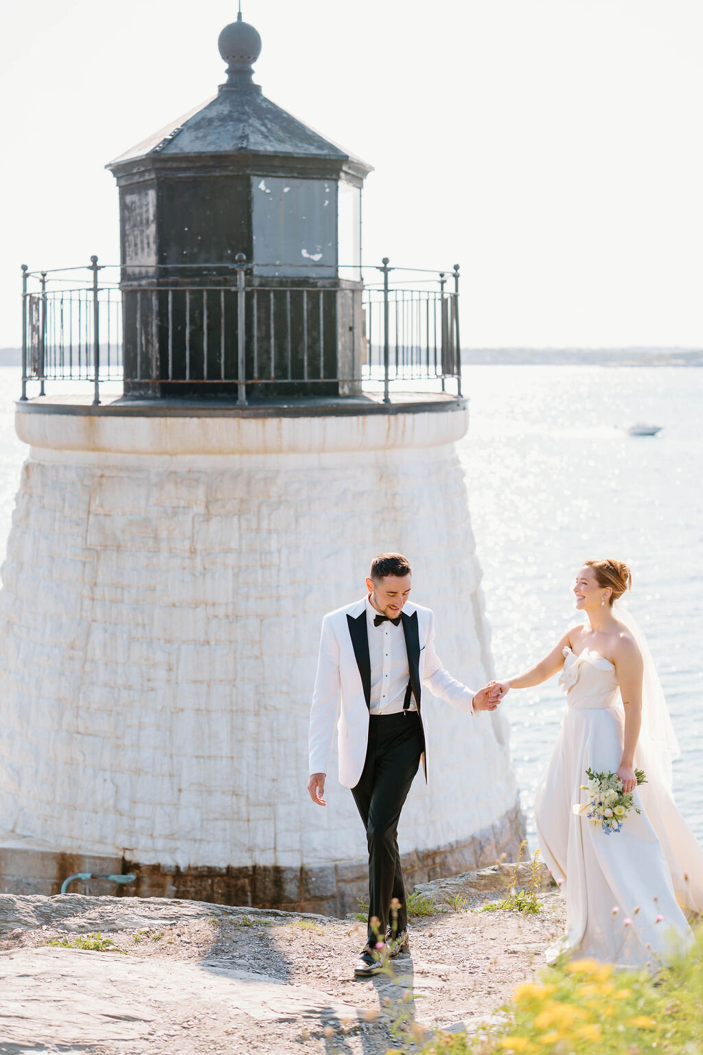 Rhode Island Elopement | A bride and groom hold hands joyfully by a lighthouse near the sea. She's in a white gown with flowers; he's in a white tuxedo jacket. 