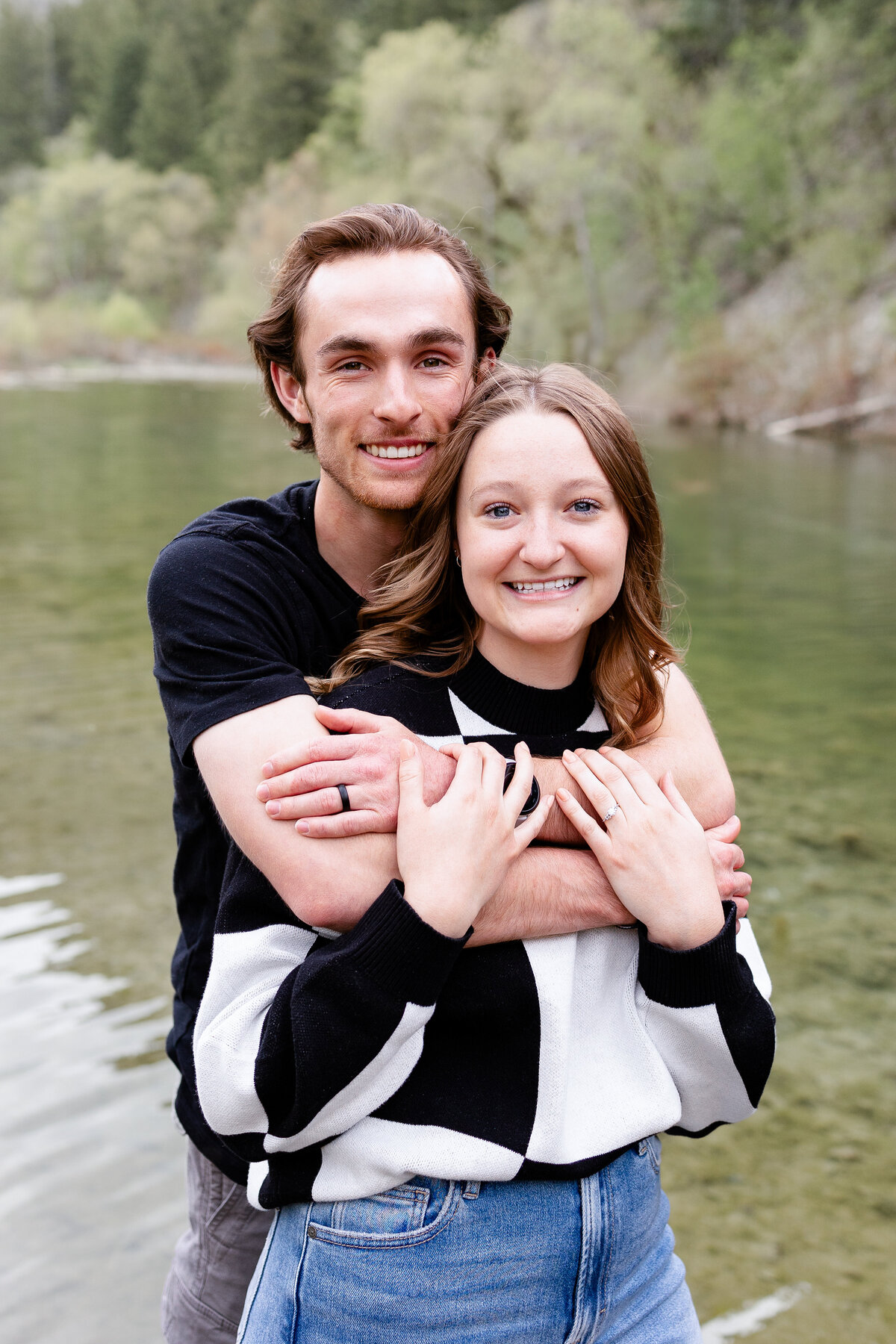 A couple standing near the water’s edge, embracing warmly with scenic mountains and trees in the background at Provo Canyon.