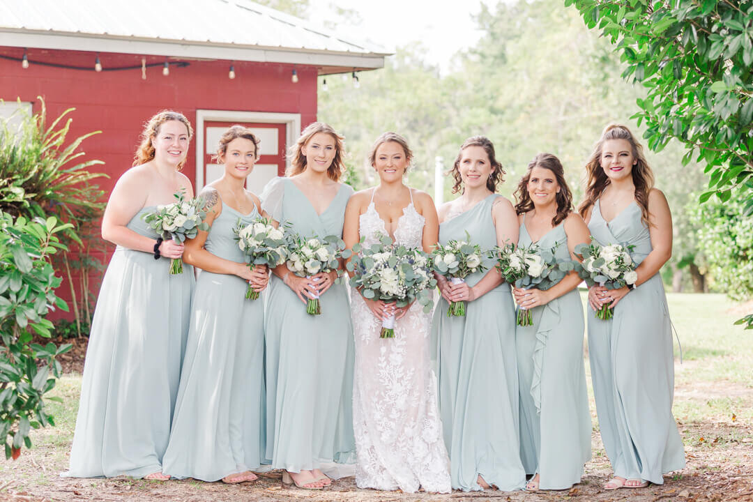 Bride stands with bridesmaids with green, white and blue flowers and blue dresses in front of a red barn.