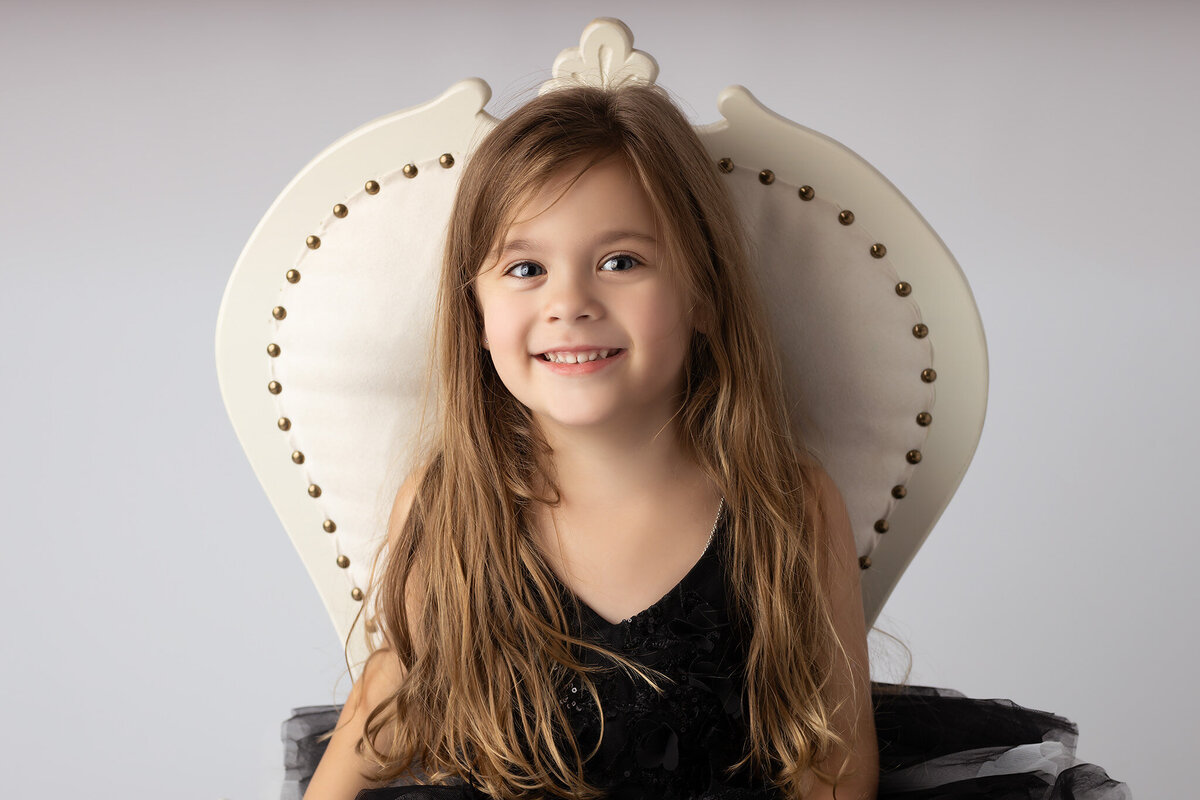 A smiling young girl in a black dress with gray ruffles, sitting comfortably in a vintage chair.