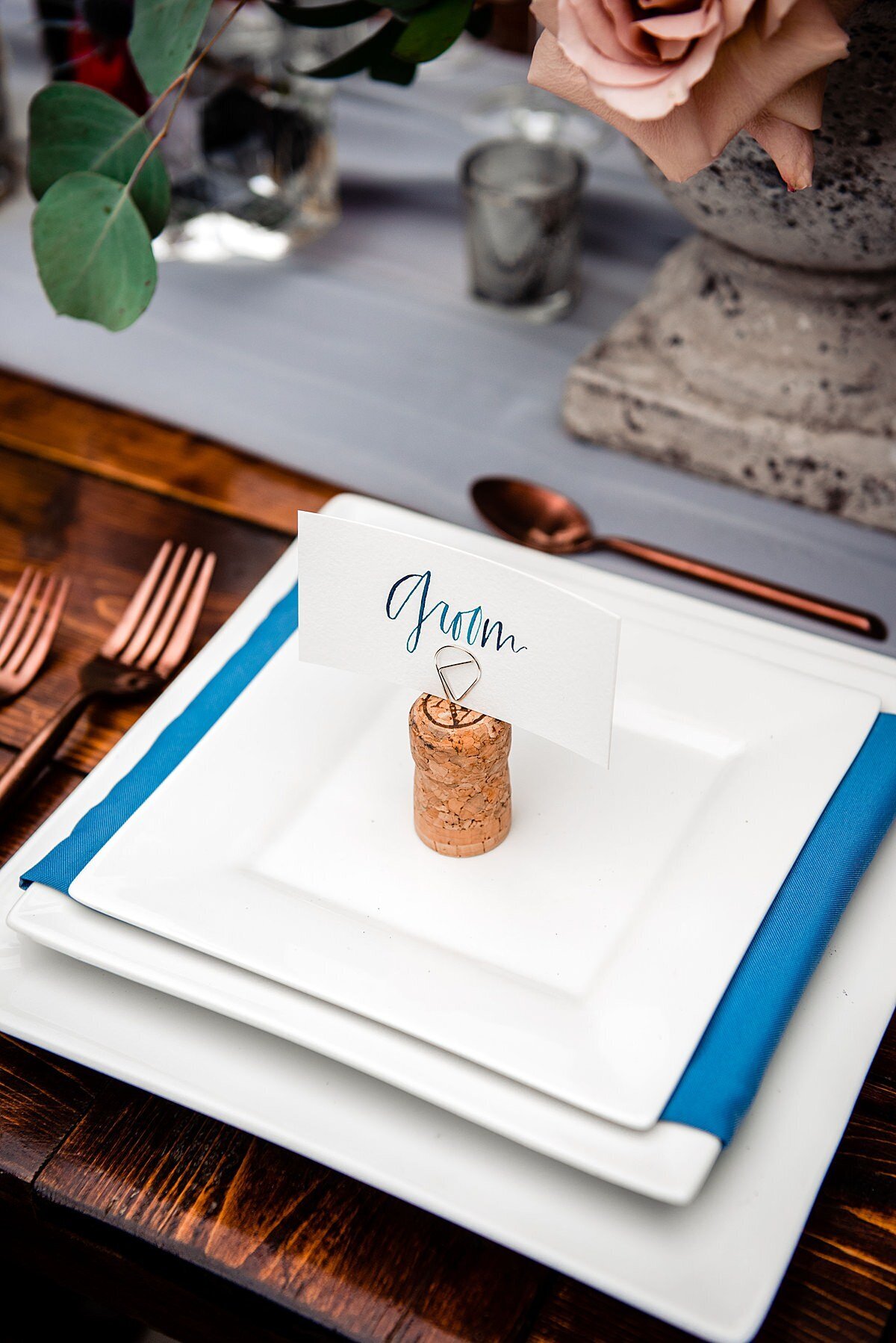 Detail of farm table set with matte copper flatware and a gray sheer organza table runner  with blush roses. The white square dinner plate is stacked with a square white salad plate wrapped in a teal blue napkin with a square white dessert plate topped with a wine cork stand holding a seating card. The seating card has Groom written in teal hand calligraphy at Arrington Vineyards.