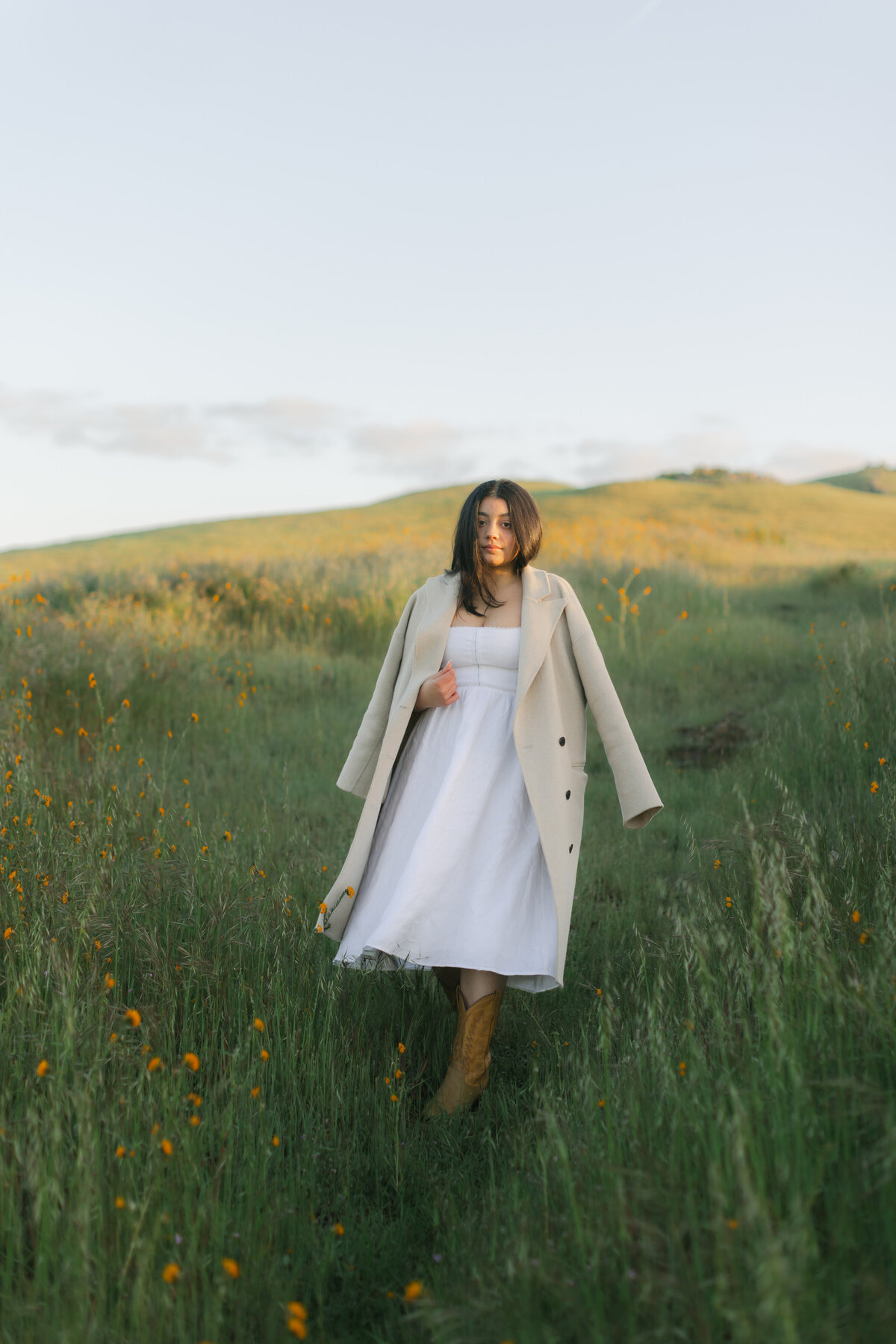 Full-body portrait of a woman in a white dress and tan coat standing in a flower-filled field in Greenville, SC. Editorial-style image by Dani Lacey with soft golden hour light.