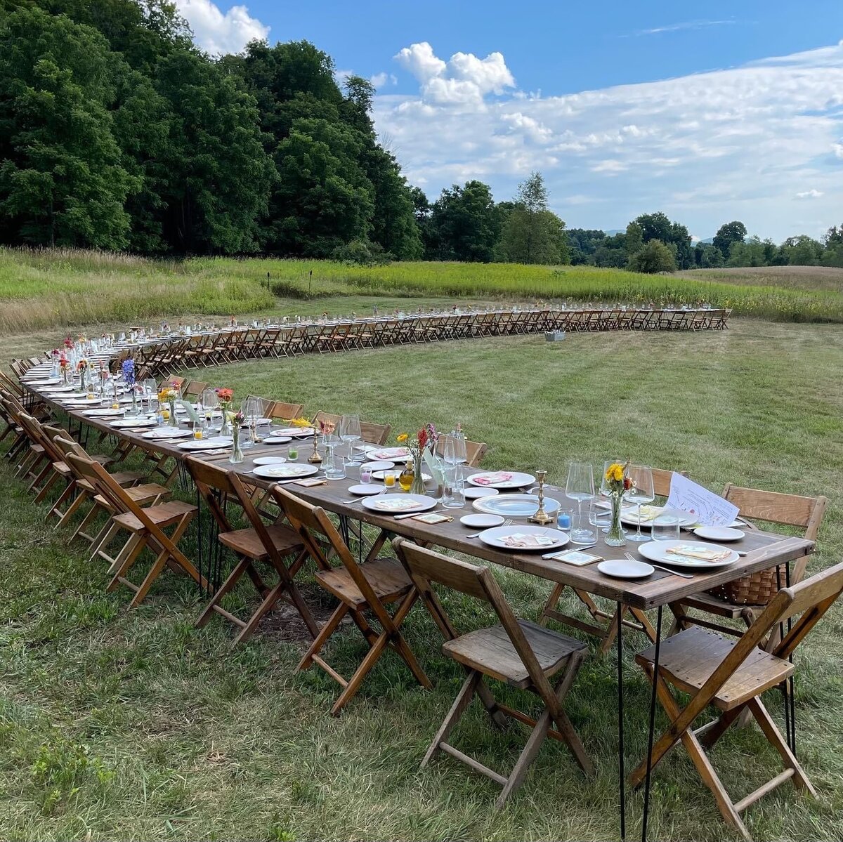 A long, curved setup of wooden tables and chairs is arranged outdoors on a grassy field. The tables are set with plates, glasses, and small flower arrangements. The scene is surrounded by greenery and trees.
