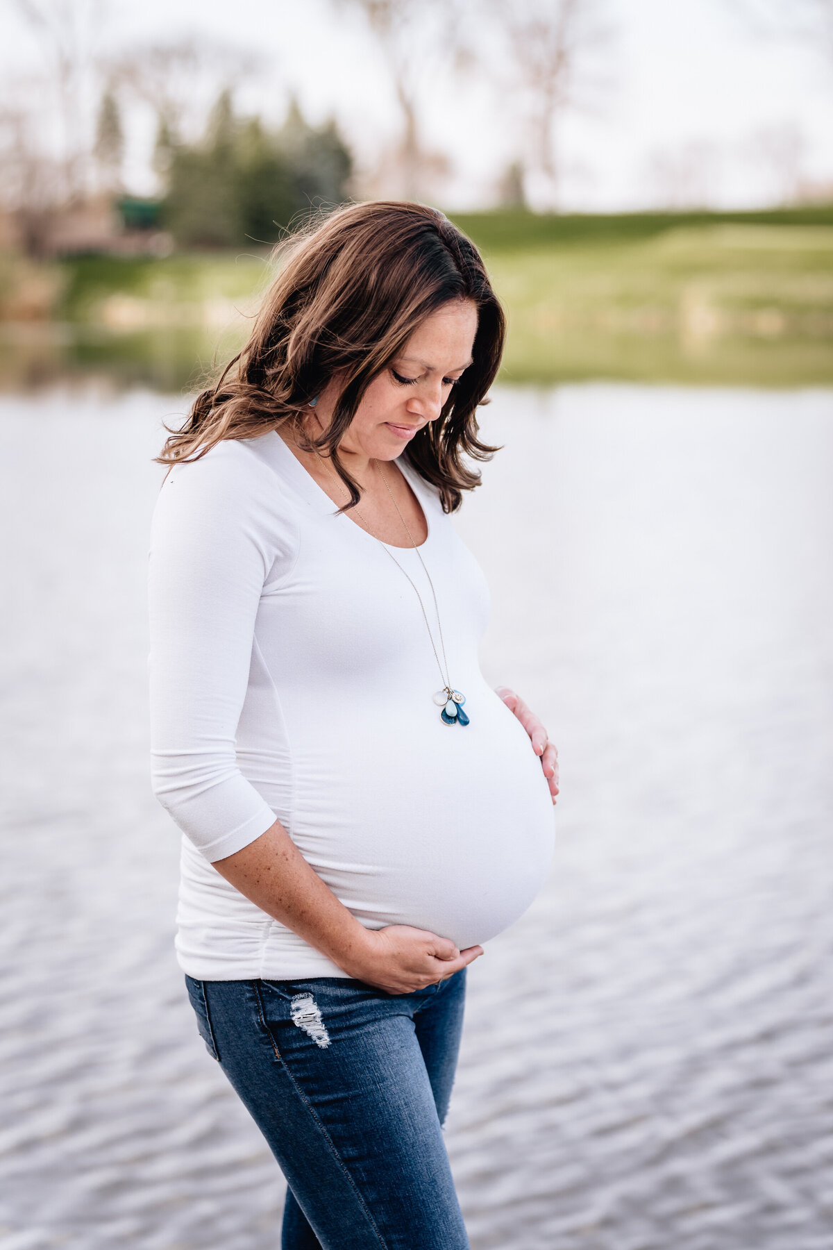 A pregnant woman stands by a calm lake, looking down at her belly with a peaceful expression, her hands gently cradling it.
