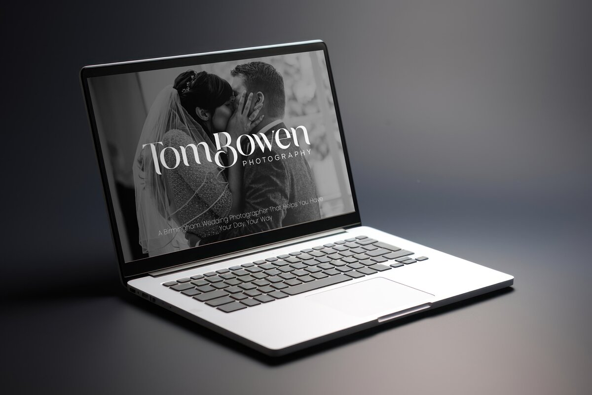 A laptop displaying the Tom Bowen Photography website, featuring a black and white image of a bride and groom kissing. The text "Tom Bowen Photography" is prominently displayed on the screen, along with the tagline "A Birmingham Wedding Photographer That Helps You Have Your Day, Your Way." The laptop is set against a dark background, highlighting the elegant and professional design of the website.