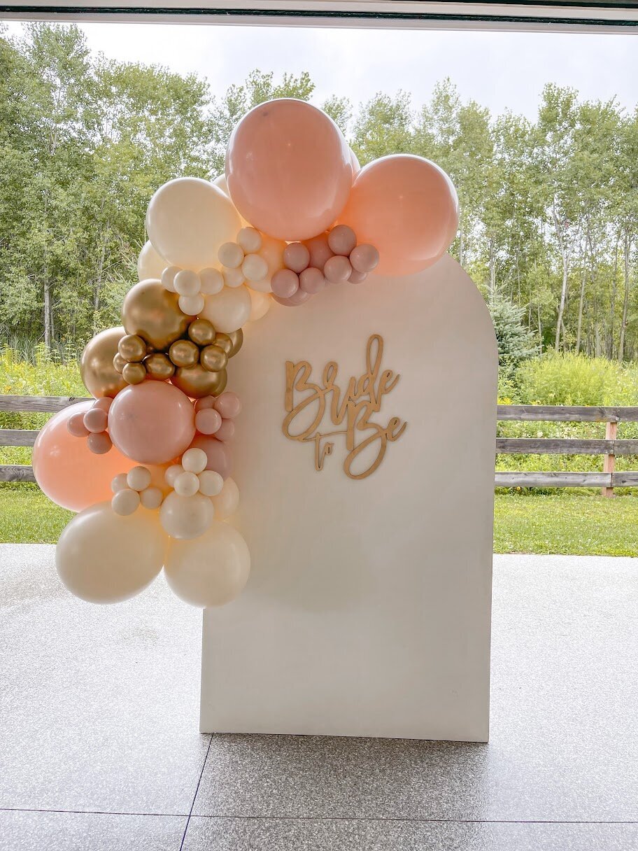 Pink, gold, and white balloons set up on a white wooden arch backdrop. A gold sign attached that says Bride to Be.