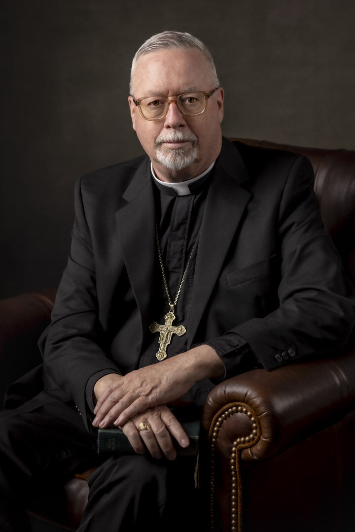 Studio portrait in CT of Arch Bishop Christopher Coyne in black suit with cross necklace, seated in leather chair by Rob Faber Photography.