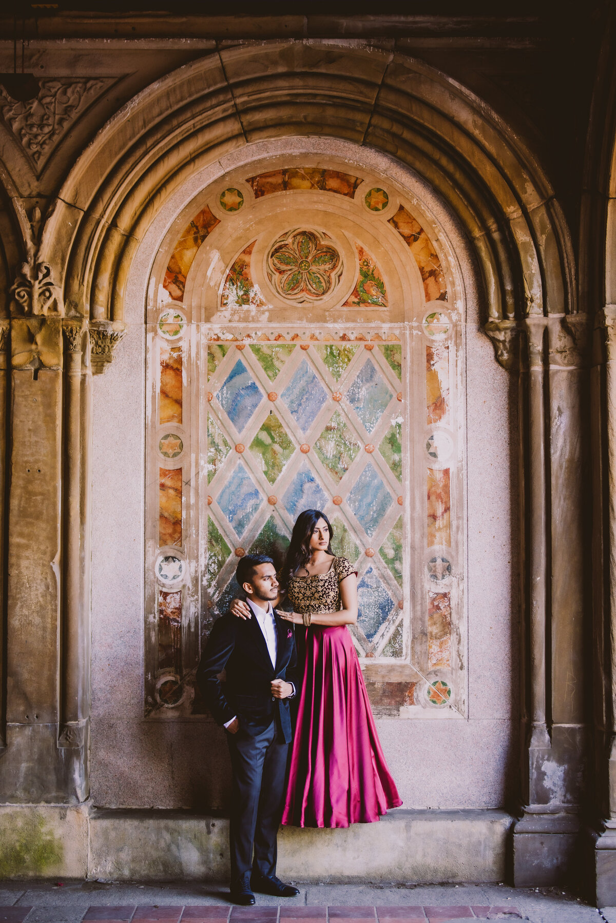 An engaged couple in traditional indian clothing and a suit standing in front of a mural looking outside the bethesda terrace arcade at central park nyc