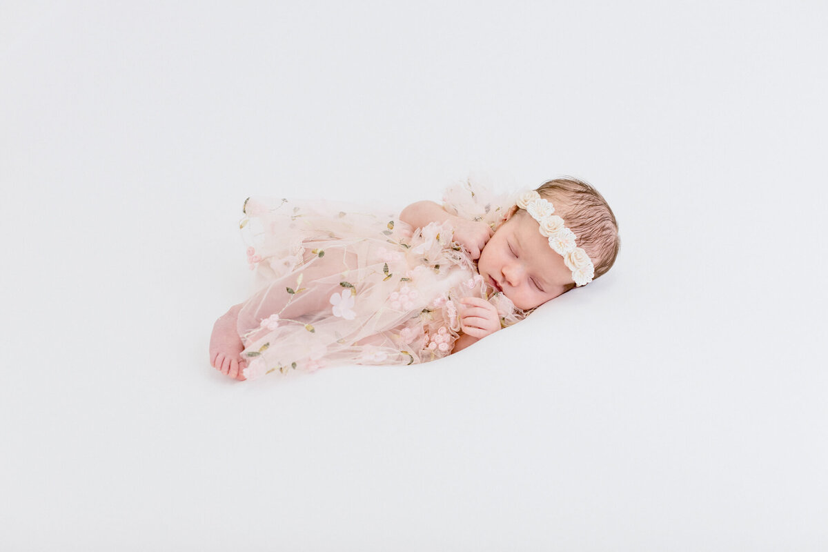 A sleeping baby is lying on a white surface, wearing a delicate floral dress and a matching headband. The baby appears peaceful and serene, surrounded by a soft, light background.