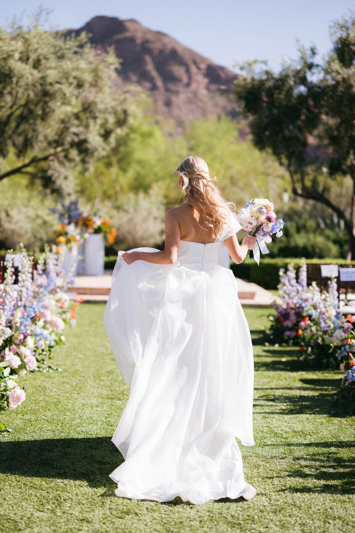 Bride walking through colorful floral aisle at El Chorro with Camelback Mountain behind her, captured by Scottsdale wedding photography team.