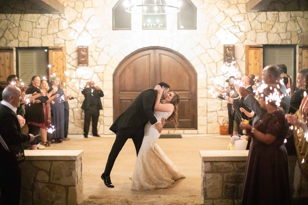 man dips bride with wood arched doors and guests with sparklers at Sendera Springs