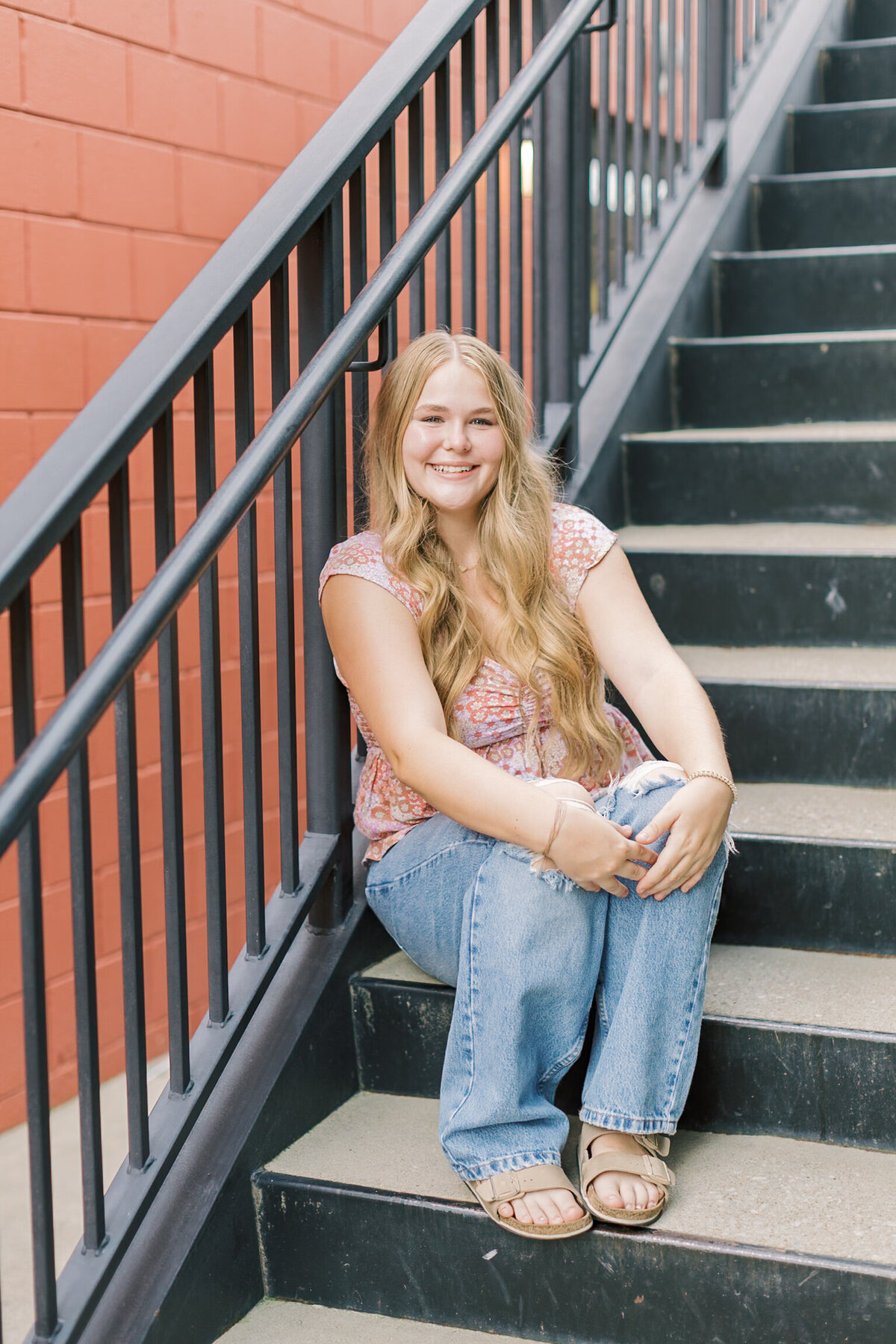A high school senior sitting on city steps, smiling confidently in casual jeans and a pink top — Portrait photography in Raleigh.