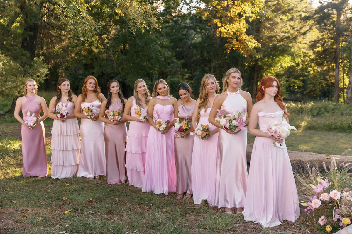 Bridesmaids standing outdoors in soft pink dresses holding petite bouquets of roses, lilies, and seasonal blooms arranged in a cohesive pastel palette
