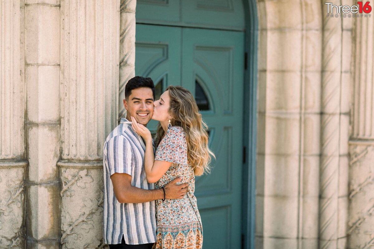 Bride to be gives her man a big kiss on the cheek as he holds her during their engagement session