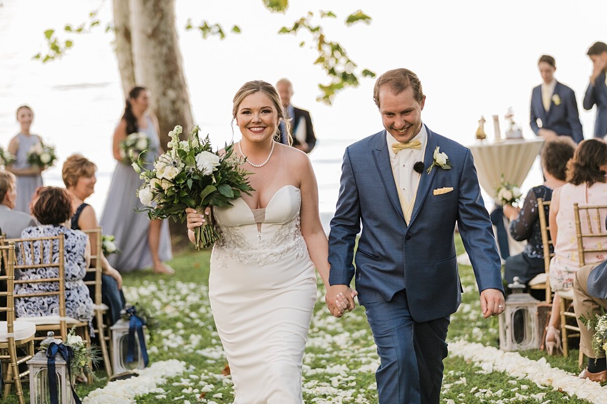 A bride wearing fitted strapless wedding dress with a plunging v neckline holds a large bouquet of greenery, qhite roses, white hydrangea and white stock. The groom, wearing a blue suit and gold vest holds the bride's hand as they walk down the aisle covered in white flower petals. Wedding guests seated in gold chivari chairs watch them leave at The Estate at Cherokee Dock.