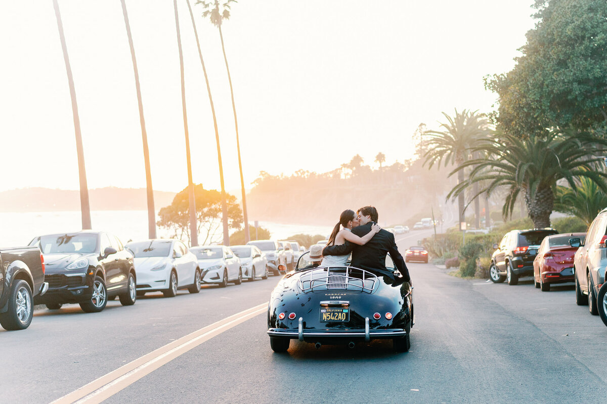 Bride and groom kiss at sunset while sitting on the back of a vintage roadster coupe while driving along Santa Barbara coastline