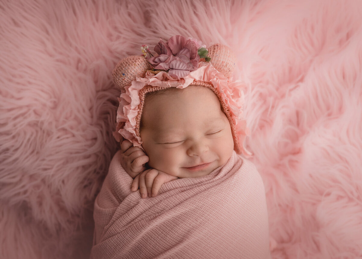 A smiling newborn baby sleeps in a pink swaddle and bonnet on a matching fur blanket in the studio of a Des Moines Newborn Photographer