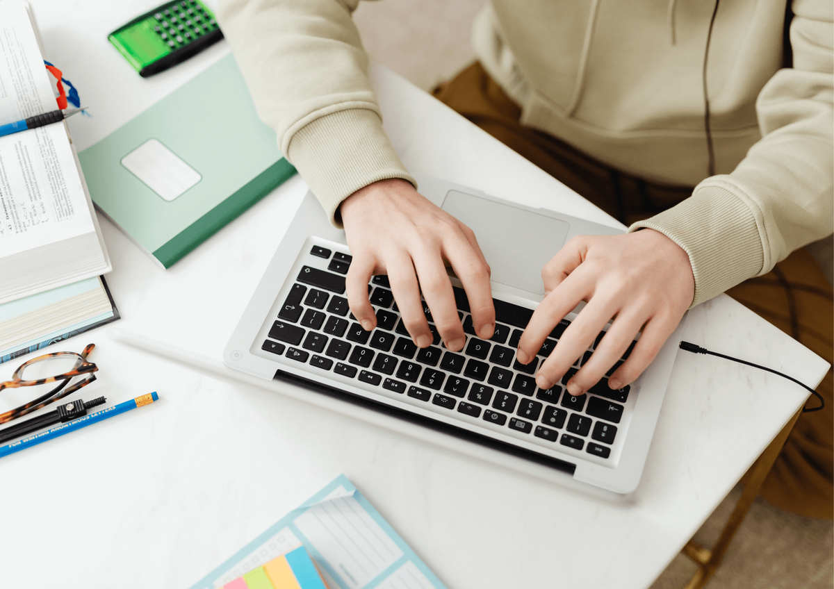 Personne tapant sur un ordinateur portable, entourée de livres, lunettes et carnet, illustrant la création de contenu digital.