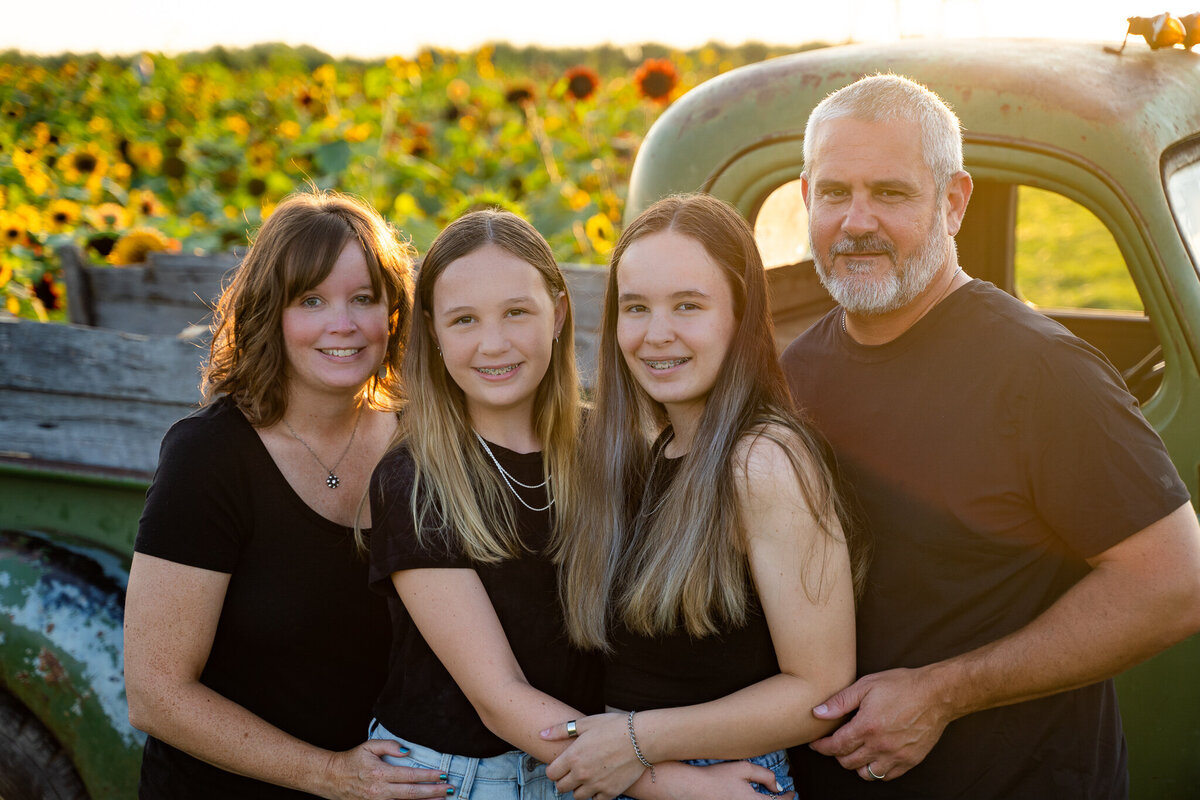 Mother and father with their teenage daughters hold each other for a family portrait in a field of sunflowers at golden hour at Lynd Fruit Farm in Pataskala, Ohio.