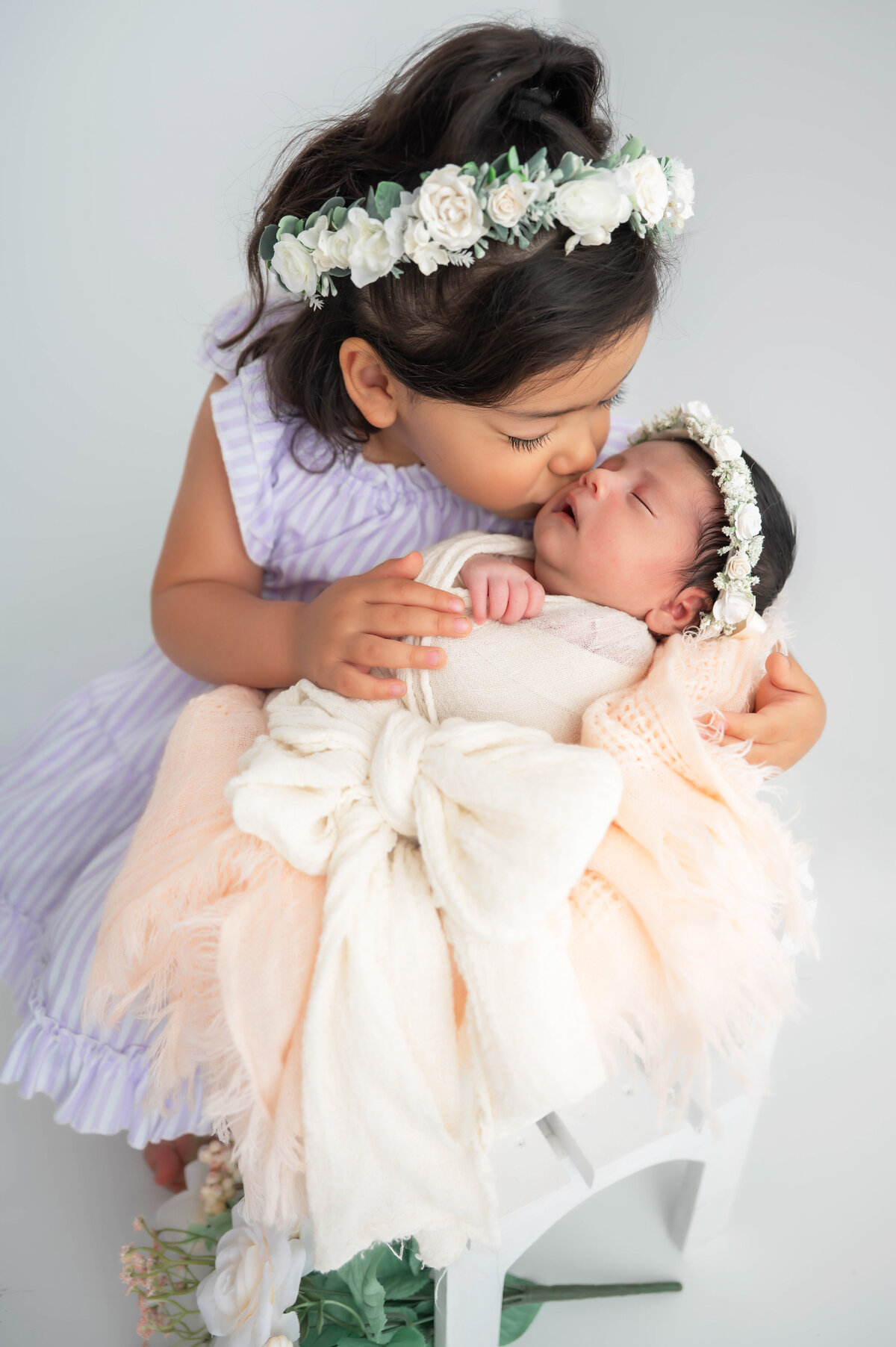 Toddler in a lilac dress gently kissing a baby wrapped in a cream bow, both wearing matching floral crowns.