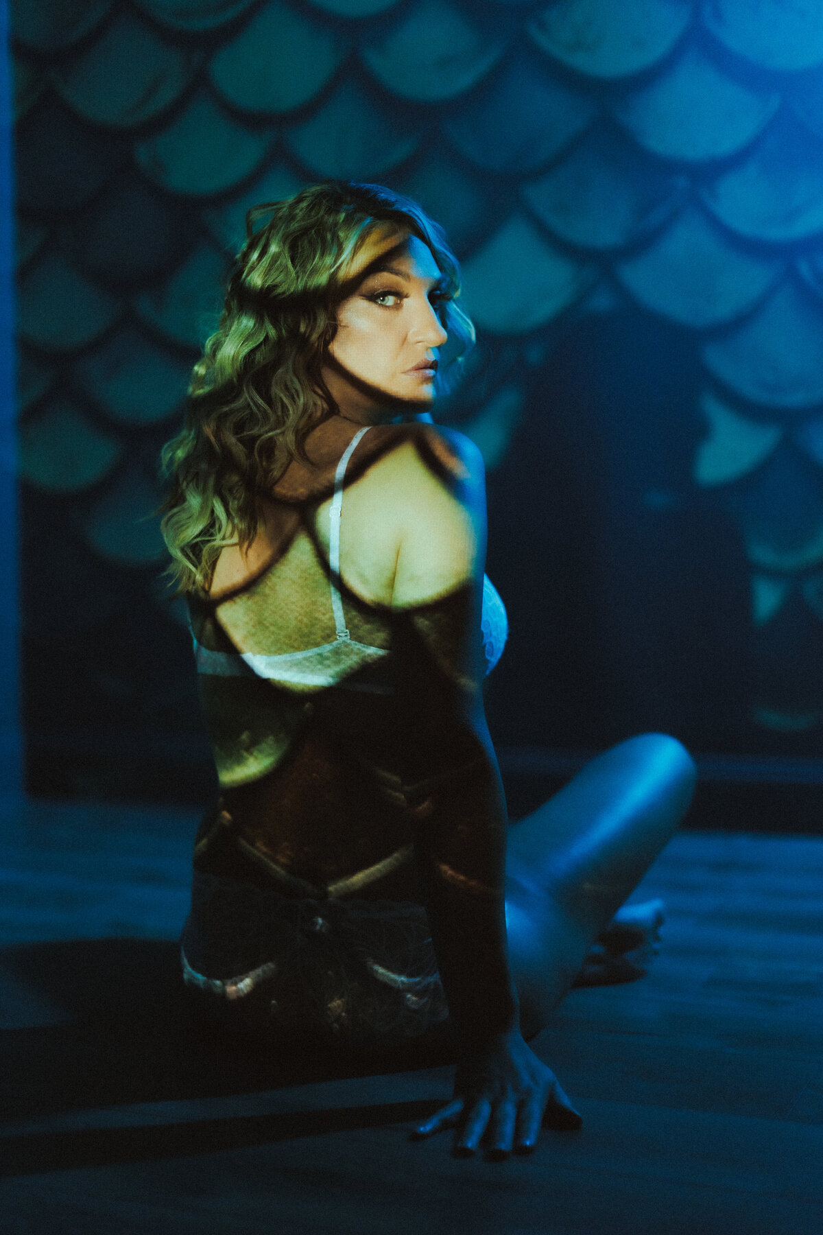 Woman in white lingerie poses with mermaid scale projections on her back in a blue-toned conceptual boudoir shoot in Tampa, Florida.