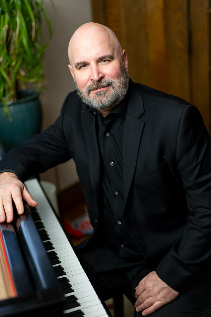 rofessional headshot of man in black shirt seated at piano smiling confidently with wood background new york.