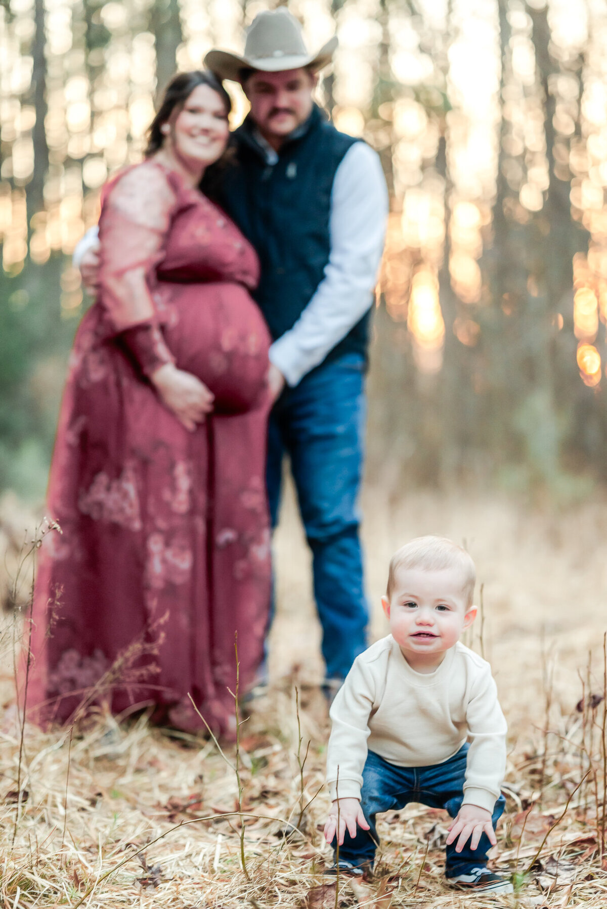 A toddler squats in the tall grass in front of his parents. The parents put their arms around each other and on mom's pregnant belly.