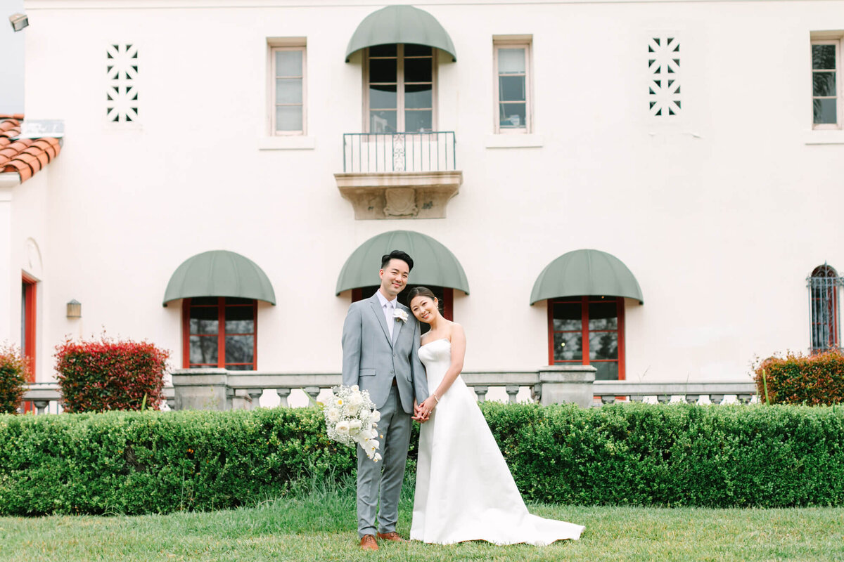 A joyful couple stands on a grassy lawn in front of an elegant building with green awnings. The bride, in a white dress, leans on the groom holding flowers.