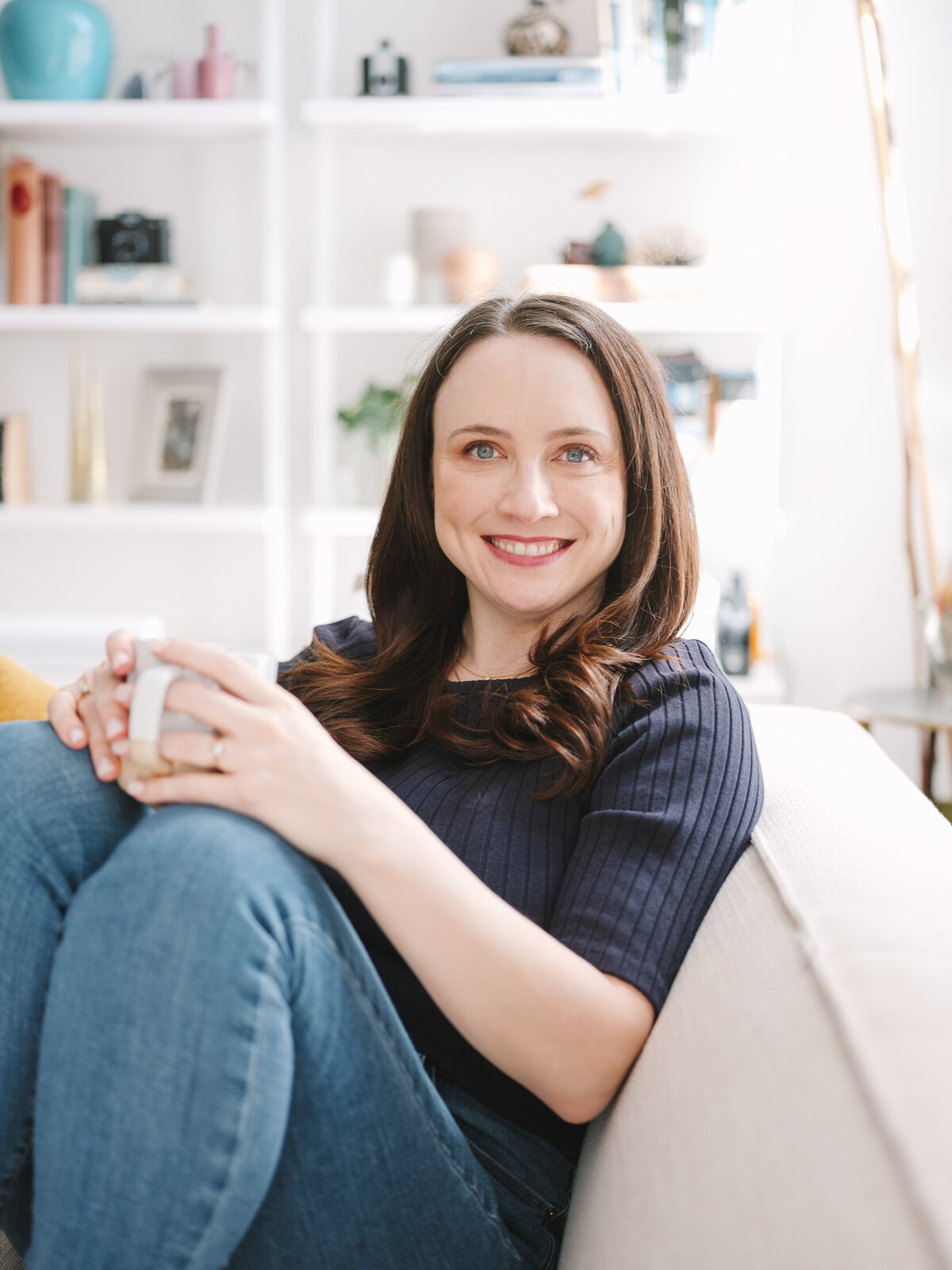 Brunette woman smiling while holding mug on couch during cozy New York City brand photo session.