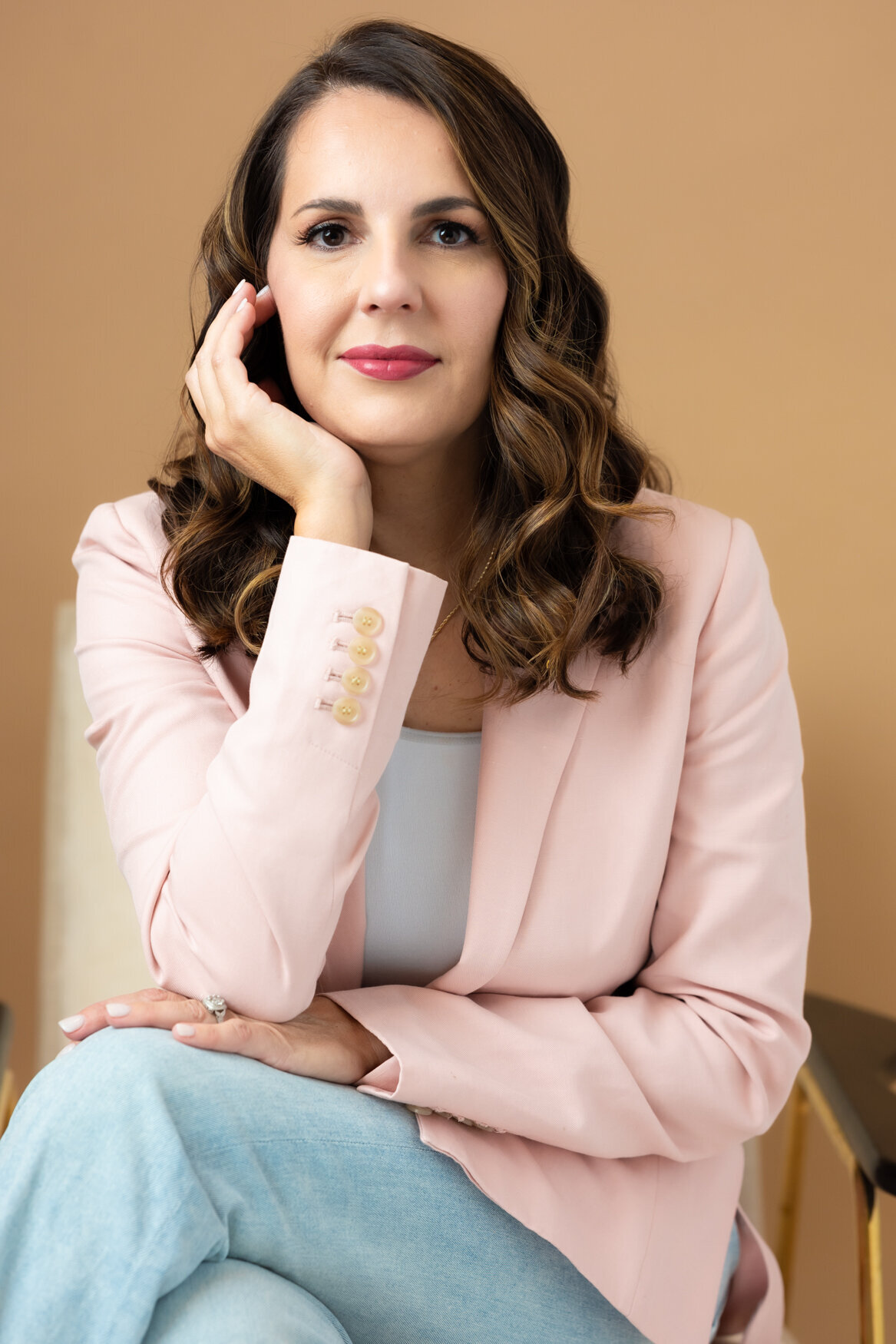A woman sitting on. a chair with her legs crossed and one hand touching her face, looking to the camera with a smile