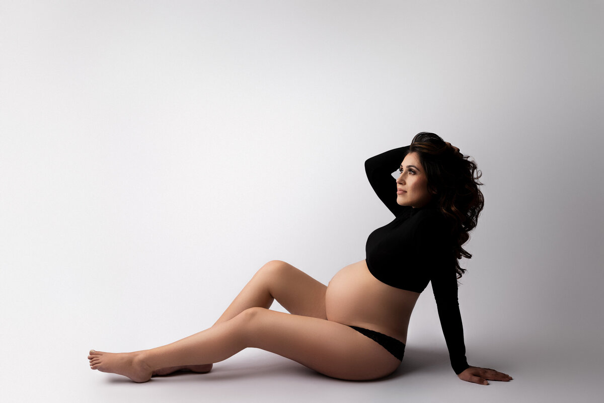 Maternity photo by Katie Marshall, New Jersey's premier maternity photographer. Woman in a long sleeve crop top and black panties sits facing left, with her weight supported on her forearm. Her left arm gently brushes hair off her face, creating an evocative and artistic pose.